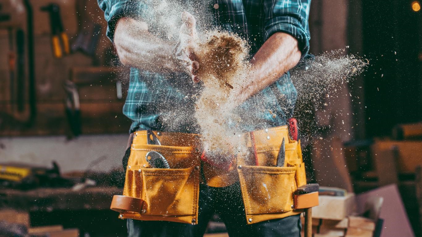 Man with a construction belt, filled with tools, fastened around his waist. His hands are clapping together, throwing construction dust into the air. 