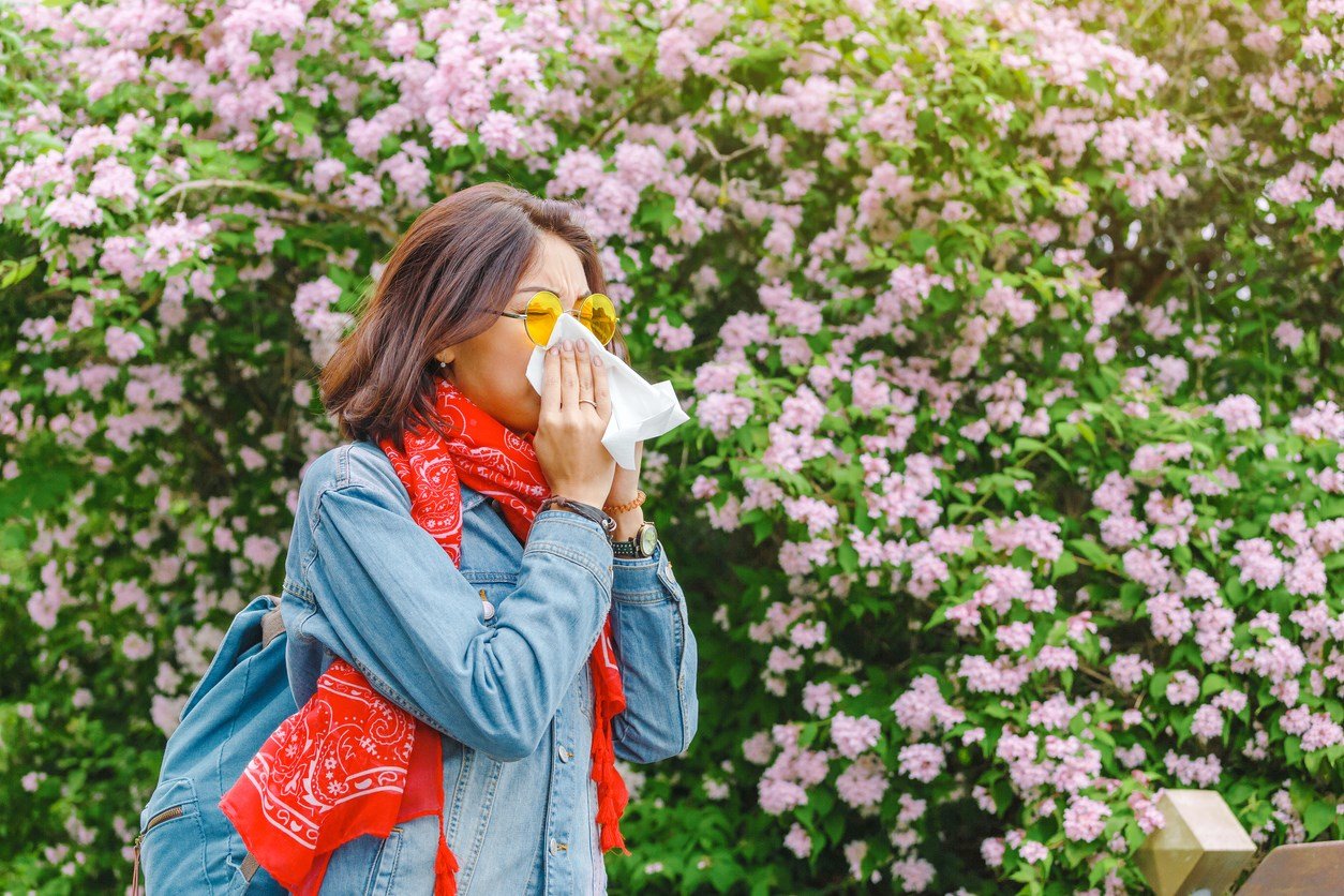 Woman blowing her nose in front of a flower bush. 