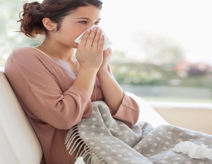 Woman sitting on the couch, wrapped in a blanket and blowing her nose into a tissue. 