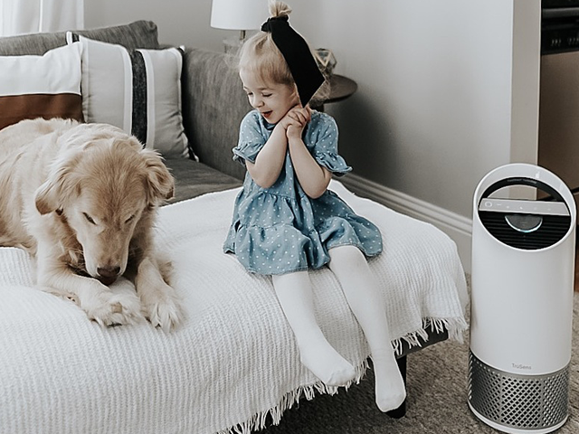 Little girl sitting on a bed smiling at her dog with a TruSens Air Purifier next to her. 