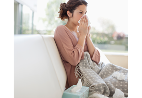 Woman sitting on the couch, blowing her nose into a tissue.